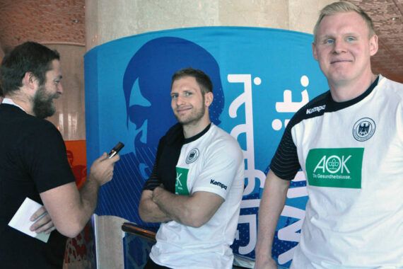 Ein Mann interviewt zwei Sportler in weiß-schwarzen Sporttrikots mit grünem AOK-Logo, die in einer Halle vor einem blauen Banner stehen. Beide Sportler lächeln, einer lehnt sich an ein Geländer.