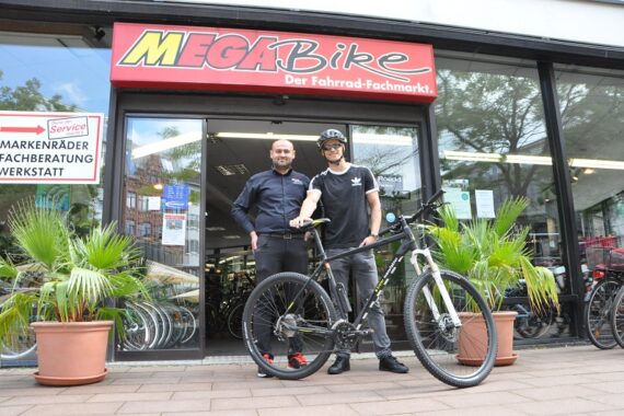 Zwei Personen stehen vor einem Fahrradgeschäft namens Mega Bike. Eine Person hält ein Mountainbike, während beide in die Kamera lächeln. Das Geschäft hat Palmen am Eingang und viele Fahrräder im Inneren.