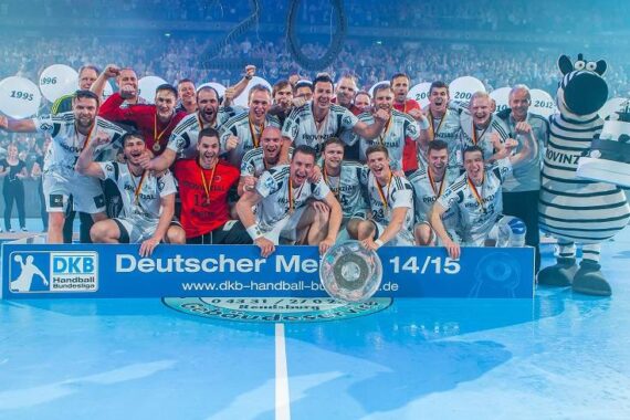 Die Handballmannschaft der Männer feiert den Gewinn der Deutschen Meisterschaft 2014/2015 und posiert mit Medaillen und einem Pokal hinter einem Banner auf einem blauen Spielfeld, mit einem Zebra-Maskottchen und jubelnden Fans im Hintergrund.