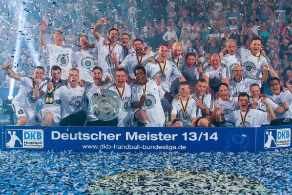 Eine jubelnde Handballmannschaft in weißen Trikots feiert den Gewinn der deutschen Meisterschaft 2013/14 und posiert mit Medaillen und einem Pokal, während um sie herum Konfetti fällt. Im Hintergrund jubelt die Menge.