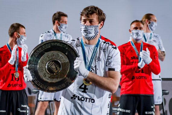 Ein Handballspieler in einem weißen Trikot hält ein großes Meisterschaftsschild, umgeben von Mannschaftskameraden in rot-weißen Trikots mit gestreiften Gesichtsmasken und Medaillen, die auf einer Bühne feiern.