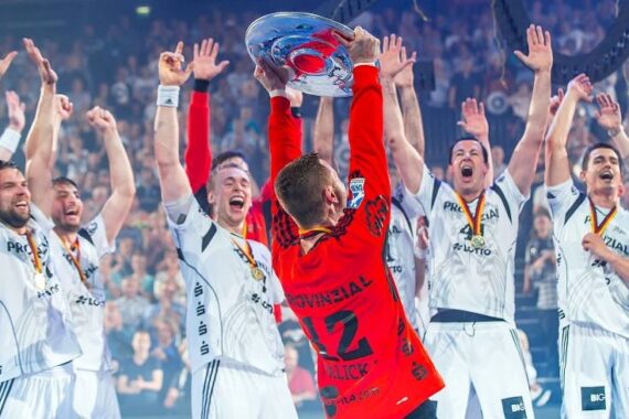 Eine Gruppe jubelnder Athleten in weißen Trikots feiert, während ein Mannschaftskamerad in einem roten Trikot eine große silberne Trophäe über seinen Kopf hebt, während Konfetti fällt und die Menge im Hintergrund jubelt.