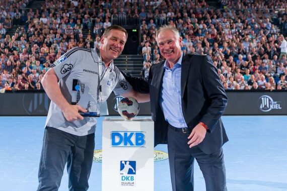 Zwei Männer lächeln für ein Foto in einer Sporthalle; der eine hält einen Pokal und einen kleinen Handball, der andere steht daneben. Sie stehen an einem Podium mit einem DKB-Logo, und im Hintergrund sind Zuschauer.