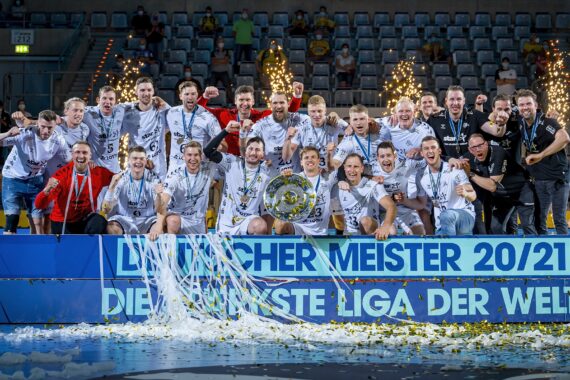 Eine jubelnde Männerhandballmannschaft in weißen Trikots feiert auf einem Podium mit Konfetti, Medaillen und einem Pokal. Im Hintergrund wird ein Feuerwerk gezündet, und auf einem blauen Banner steht "LIQUI MOLY HBL DEUTSCHER MEISTER 20/21".