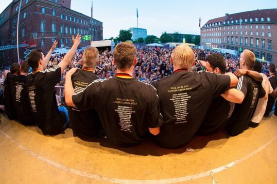 Eine Gruppe von Menschen in passenden schwarzen Hemden sitzt auf einer Bühne, die Arme umeinander gelegt, und blickt auf eine große jubelnde Menge bei einer Veranstaltung im Freien auf einem Stadtplatz.