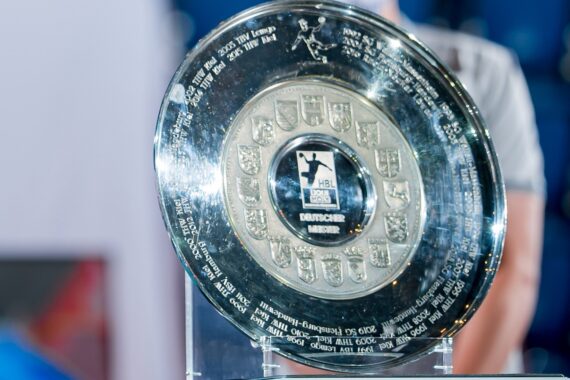 Eine Nahaufnahme des Meisterschaftsschildes der Handball-Bundesliga, einer großen, glänzenden silbernen Trophäe mit eingravierten Namen, Logos und Schildern, die auf einem Glasständer mit unscharfem Hintergrund steht.