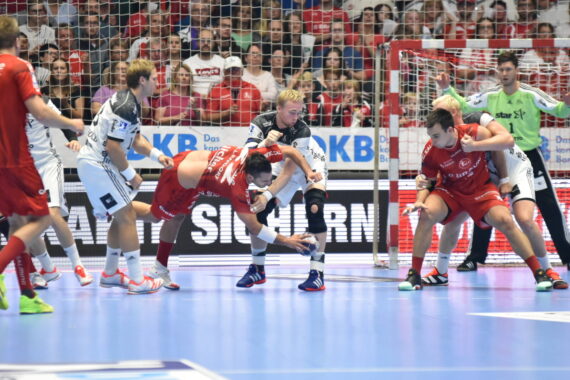 Mehrere männliche Handballspieler in rot-weißen Trikots wetteifern in der Nähe des Tores. Ein Spieler in Rot beugt sich vor, um den Ball aufzuheben, während andere den Ball verteidigen und ein Torwart im Hintergrund zuschaut.