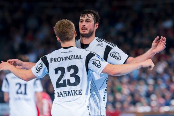 Zwei männliche Handballspieler in weiß-schwarzen Trikots mit "PROVINZIAL" und "DAHMKE 23" auf dem Rücken feiern auf dem Spielfeld und strecken sich in einer überfüllten Arena die Arme entgegen.