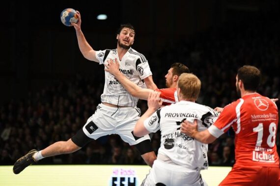 Ein männlicher Handballspieler in einem weißen Trikot springt in der Luft, um den Ball zu werfen. Er ist von Verteidigern in roten Trikots umgeben, während eines wettbewerbsorientierten Hallenspiels mit einer Menschenmenge im Hintergrund.