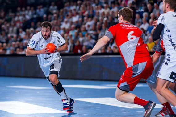 Ein Handballspieler in einem weißen Trikot läuft mit einem orangefarbenen Ball, während er von Spielern in roten Trikots während eines Spiels in einer überfüllten Halle verfolgt wird.