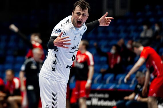 Ein männlicher Handballspieler in einem weißen Trikot schreit und gestikuliert mit ausgestreckten Armen während eines Spiels, während im Hintergrund andere Spieler und Trainer in roten Trikots zu sehen sind.
