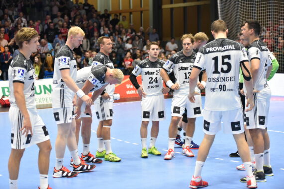 Eine Herren-Handballmannschaft in weißen Trikots mit schwarzen Akzenten versammelt sich auf einem Hallenplatz und scheint während eines Spiels Taktiken zu besprechen. Die Zuschauer füllen die Tribünen im Hintergrund.