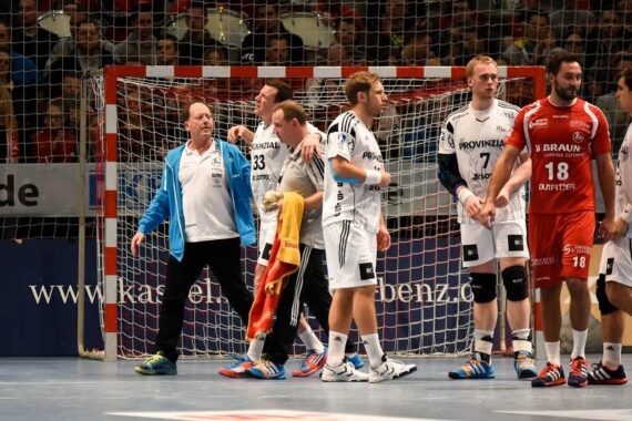 Eine Gruppe männlicher Handballspieler in Weiß und eine in Rot stehen zusammen auf einem Hallenplatz in der Nähe des Tores, während ein Trainer oder ein Mitarbeiter in der Nähe gestikuliert. Die Zuschauer schauen von den Tribünen im Hintergrund zu.
