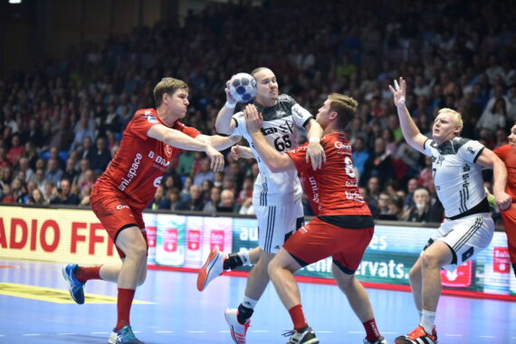 Vier männliche Handballspieler sind während eines Spiels in Aktion, wobei zwei in roten Trikots versuchen, einen Spieler in einem weißen Trikot, der den Ball hält, zu blockieren, während ein weiterer Spieler in einem weißen Trikot im Hintergrund zu sehen ist. Die Arena ist mit Zuschauern gefüllt.