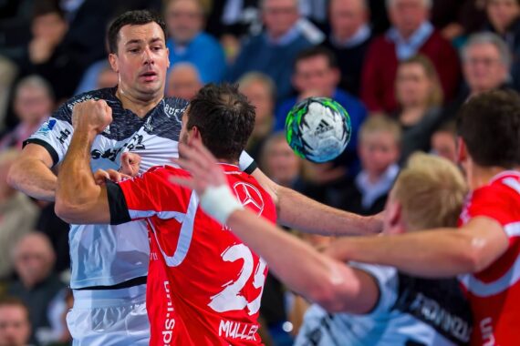Drei männliche Handballspieler kämpfen während eines Spiels um den Ball, einer in einem weißen und zwei in einem roten Trikot. Das Spiel ist intensiv und die Zuschauer im Hintergrund sind unscharf.