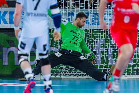 Ein Handballtorwart in grünem Trikot und schwarzer Hose streckt sich, um einen Schuss vor dem Tor abzuwehren, während sich Spieler in weißen und roten Trikots um ihn herum auf dem Spielfeld bewegen.