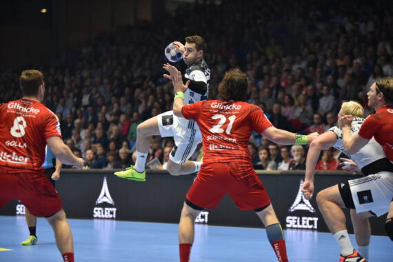 Ein Handballspieler in einem weiß-schwarzen Trikot springt, um den Ball zu werfen, während drei Spieler in roten Trikots ihn abwehren, während ein großes Publikum im Hintergrund zusieht.