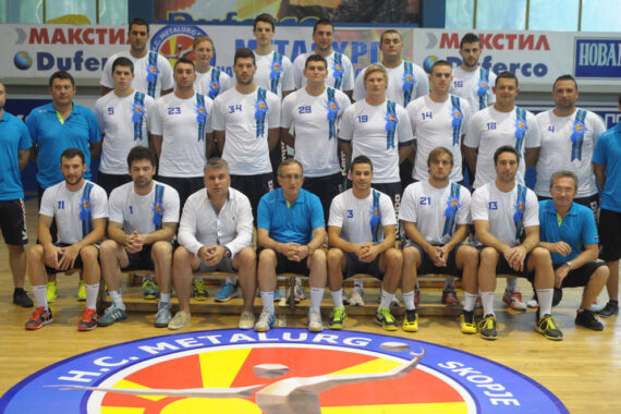 Eine Handballmannschaft der Männer, H.C. Metalurg Skopje, stellt sich in einer Halle in zwei Reihen mit Spielern in weißen und Trainern in blauen Hemden auf einem Spielfeld mit dem Logo und dem Namen der Mannschaft auf.