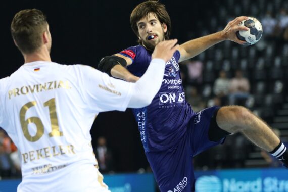 Ein Handballspieler in einem blauen Trikot springt und macht sich bereit, den Ball zu werfen, während ein Gegner in einem weißen Trikot mit der Nummer 61 einen Arm hebt, um ihn zu blockieren. Im Hintergrund sind leere Plätze und unscharfe Zuschauer zu sehen.