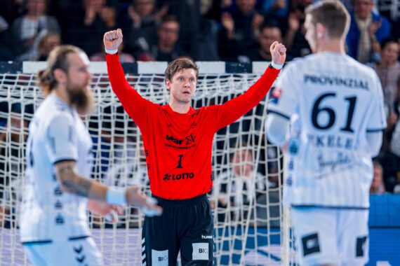 Ein Handballtorhüter in einem roten Trikot reißt vor dem Tor jubelnd beide Arme hoch, während zwei gegnerische Spieler in weißen Trikots während eines Spiels in der Nähe des Spielfelds stehen.