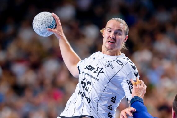 Ein männlicher Handballspieler in einem weißen Trikot springt mit einem Ball in der Hand und bereitet sich auf einen Wurf vor, während ein Gegner in Blau versucht, ihn zu blockieren; im Hintergrund ist eine verschwommene Menschenmenge zu sehen.