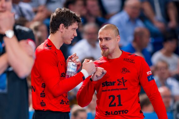 Zwei männliche Sportler in roten Trikots, von denen einer eine Wasserflasche in der Hand hält, stoßen sich am Rande eines Handballspiels mit den Fäusten an, während im Hintergrund eine unscharfe Menschenmenge zu sehen ist.