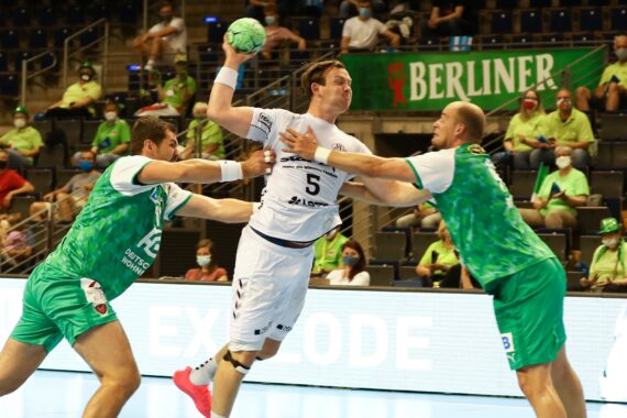 Ein Handballspieler in einem weißen Trikot springt zum Schuss, während zwei Verteidiger in Grün versuchen, ihn während eines Spiels zu blockieren. Maskierte Zuschauer schauen von der Tribüne aus zu, und im Hintergrund ist eine Berliner Fahne zu sehen.