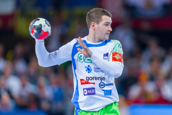 Ein männlicher Handballspieler in einem weiß-blauen Trikot bereitet sich darauf vor, während eines Spiels einen Ball zu werfen. Der Hintergrund ist mit Zuschauern unscharf.