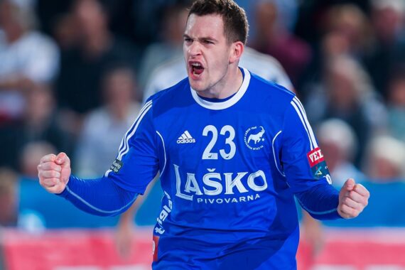 Ein männlicher Handballspieler, der ein blaues Trikot mit der Nummer 23 trägt, ballt beide Fäuste und schreit leidenschaftlich, während er ein Spiel feiert. Im Hintergrund ist eine unscharfe Menschenmenge zu sehen.