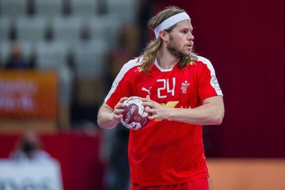 Ein männlicher Handballspieler in einem roten Trikot mit der Nummer 24 hält einen Handball und bereitet sich auf den Wurf vor. Er hat lange blonde Haare, ein weißes Stirnband und ist auf das Spiel konzentriert. Der Hintergrund ist eine Sporthalle.