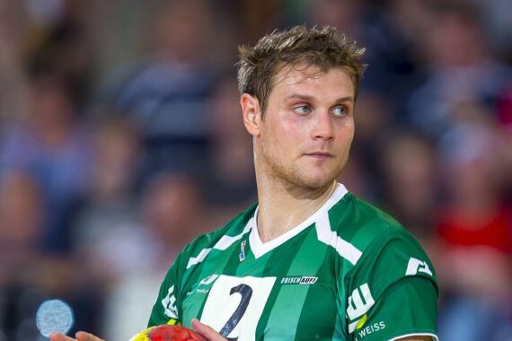 Ein männlicher Handballspieler in einem grün-weißen Trikot mit der Nummer 2 bereitet sich darauf vor, während eines Spiels einen Ball zu werfen, und blickt dabei konzentriert nach rechts. Der Hintergrund ist unscharf mit Zuschauern.