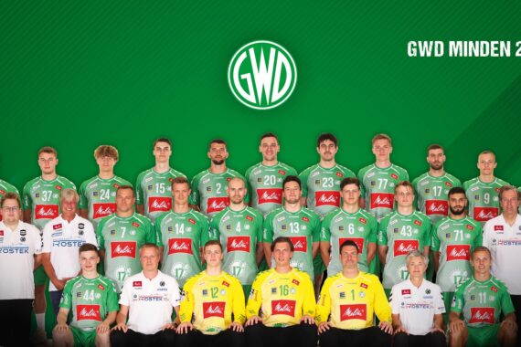Die Handballmannschaft 2021/2022 von GWD Minden posiert in Reihen vor einem grünen Hintergrund und trägt grün-weiße Trikots. Trainer und Betreuer in weißen Hemden stehen auf beiden Seiten der Spieler. Das Logo und der Name der Mannschaft sind oben zu sehen.
