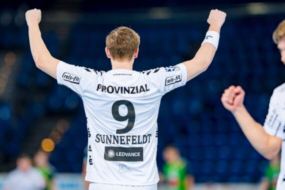 Ein Handballspieler, der ein weißes Trikot mit der Nummer 9 und dem Namen Sunnefeldt trägt, reißt auf einem Spielfeld jubelnd beide Arme in die Höhe. Andere Spieler sind im unscharfen Hintergrund zu sehen.