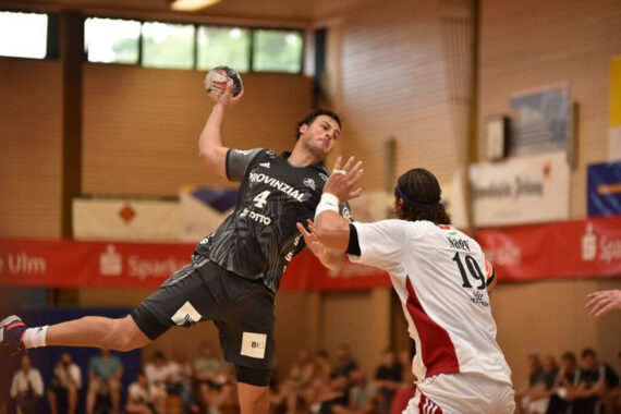 Ein Handballspieler in einem schwarzen Trikot springt, um den Ball zu werfen, während ein Abwehrspieler in einem weiß-roten Trikot versucht, ihn während eines Hallenspiels zu blockieren.