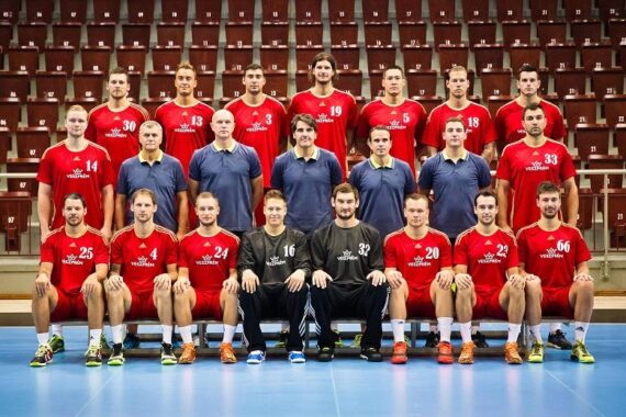 Eine Herren-Handballmannschaft stellt sich für ein Gruppenfoto in einer Sporthalle auf. Die Spieler tragen rote Trikots und sitzen oder stehen in Reihen, hinter ihnen die Trainer oder Betreuer in blauen Hemden. Im Hintergrund sind leere Tribünenplätze zu sehen.