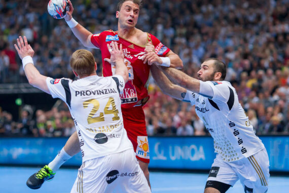 Drei männliche Handballspieler treten während eines Spiels gegeneinander an. Ein Spieler in einem roten Trikot springt, um den Ball zu werfen, während zwei Spieler in weißen Trikots mit erhobenen Armen versuchen, ihn zu blockieren. Im Hintergrund sieht eine Menschenmenge zu.