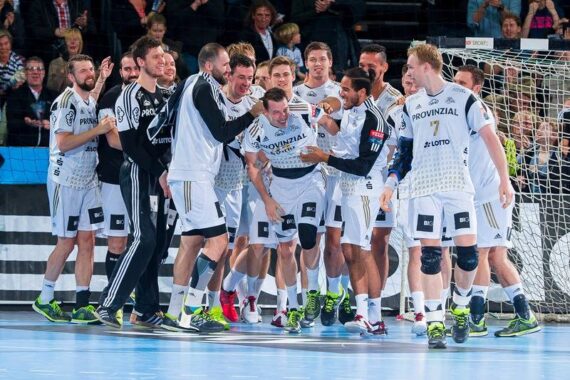 Eine Gruppe männlicher Handballspieler in weißen Trikots feiert gemeinsam auf einem Hallenplatz, lächelnd und jubelnd in der Nähe des Tores, während im Hintergrund eine Menschenmenge zusieht.