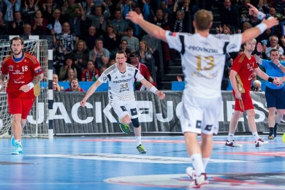 Ein Handballspieler in einem weißen Trikot feiert auf dem Spielfeld mit ausgebreiteten Armen, während im Hintergrund andere Spieler und eine jubelnde Menge zu sehen sind. Die Atmosphäre ist energiegeladen und intensiv.