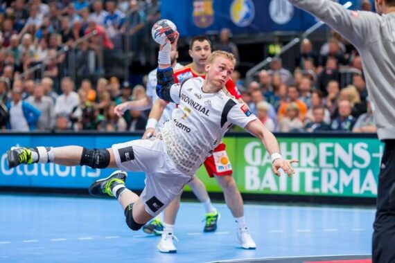Ein Handballspieler in einem weißen Trikot springt in der Luft zur Seite, um den Ball zu werfen. In der Nähe befinden sich gegnerische Spieler und ein Schiedsrichter, während im Hintergrund eine Menschenmenge zuschaut. Die Szene findet in einer Halle statt.