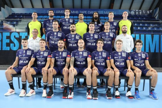 Eine Herrenhandballmannschaft in lila Trikots sowie Trainer und Betreuer in weißen und gelben Hemden stellen sich in einer Sporthalle für ein Gruppenfoto auf.