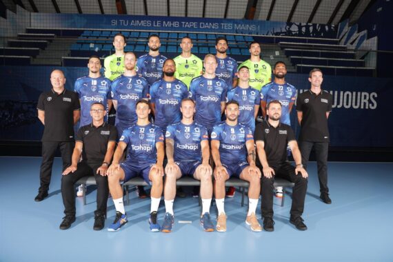 Eine Handballmannschaft der Männer stellt sich in blauen Trikots mit schwarz gekleideten Trainern in einer Sporthalle in Reihen auf. An der Wand hinter ihnen ist ein Slogan in französischer Sprache zu sehen.
