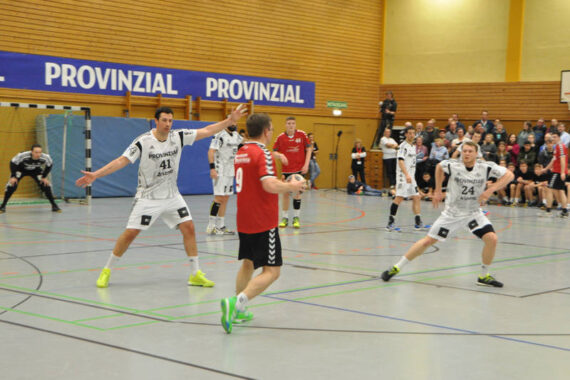 Ein Handballspiel in einer Turnhalle: Ein Spieler im roten Trikot bereitet sich auf den Wurf vor, während zwei Spieler in weißen Trikots den Ball verteidigen, während die Zuschauer im Hintergrund von der Tribüne aus zusehen.
