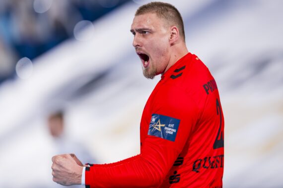 Ein männlicher Handballspieler in einem roten Trikot schreit leidenschaftlich mit geballter Faust, wahrscheinlich beim Feiern während eines Spiels. Der Hintergrund ist unscharf und lenkt die Aufmerksamkeit auf seinen Ausdruck und seine Geste.