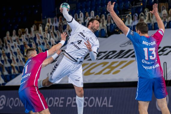 Ein Handballspieler in einem weißen Trikot springt zum Schuss, während zwei Verteidiger in blauen und rosafarbenen Trikots versuchen, ihn während eines Hallenspiels zu blockieren. Die Tribünen im Hintergrund sind mit Zuschauern besetzt.