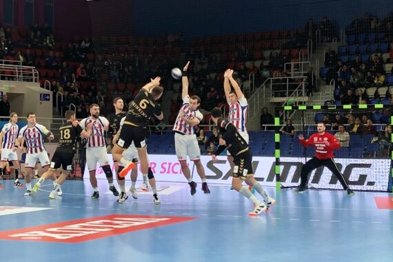 Bei einem Handballspiel springt ein Spieler in einem schwarzen Trikot auf, um den Ball zu schießen, während die Verteidiger in weiß-blau gestreiften Trikots aufspringen, um ihn abzuwehren. Der Torwart steht wachsam vor dem Tor. Zuschauer füllen die Plätze im Hintergrund.