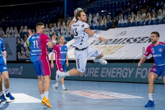 Ein Handballspieler in einem weißen Trikot springt in der Luft, um den Ball zu werfen, während die gegnerischen Spieler in rosa und blauen Trikots sich auf die Verteidigung während eines Hallenspiels vorbereiten. Im Hintergrund sind Zuschauer und leere Sitze zu sehen.