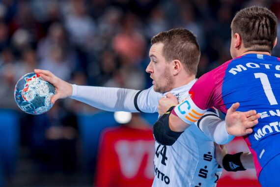 Ein Handballspieler in einem weißen Trikot streckt den Arm aus, um den Ball zu passen, während er von einem Gegner in einem blau-rosa Trikot in einem intensiven Spiel verteidigt wird.