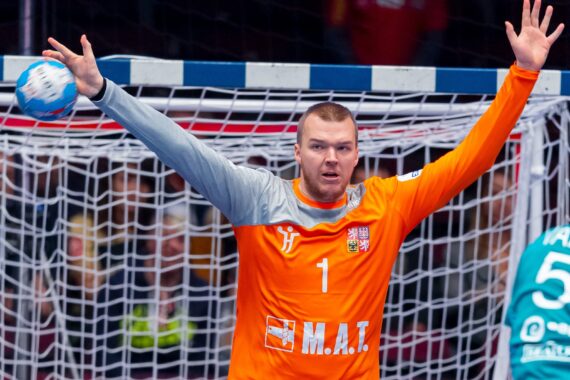 Ein Handballtorwart in einem orangefarbenen Trikot steht mit erhobenen Armen vor dem Tor und wirkt konzentriert, während sich ein Spieler in einem blaugrünen Trikot mit der Nummer 55 nähert.
