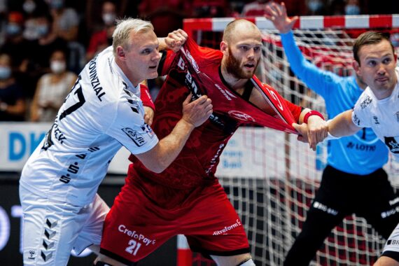 Zwei Handballspieler in weißen Trikots verteidigen sich gegen einen Spieler in einem roten Trikot, wobei einer das Trikot des roten Spielers festhält. Die Spieler wirken intensiv und konzentriert bei einem rasanten Hallenspiel. Im Hintergrund ist ein Offizieller zu sehen.