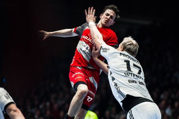 Ein Handballspieler in einem roten Trikot springt und versucht, den Ball zu schießen, während ein Spieler in einem weißen Trikot versucht, ihn während eines intensiven Spiels zu blockieren. Die Szene fängt die Action in der Luft und den Körperkontakt ein.